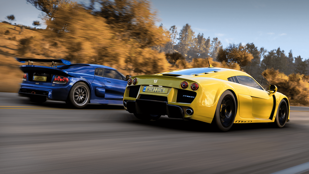 Blue and yellow Noble supercars race down the road surrounded by the glint of Autumn trees and foliage.