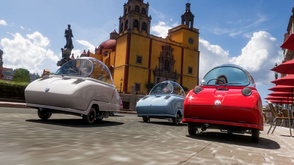 A trio of Peel Tridents, one red, one white and one blue, drive along the city streets of Guanajuato. A statue, orange architectural building, and red table umbrellas can be seen.