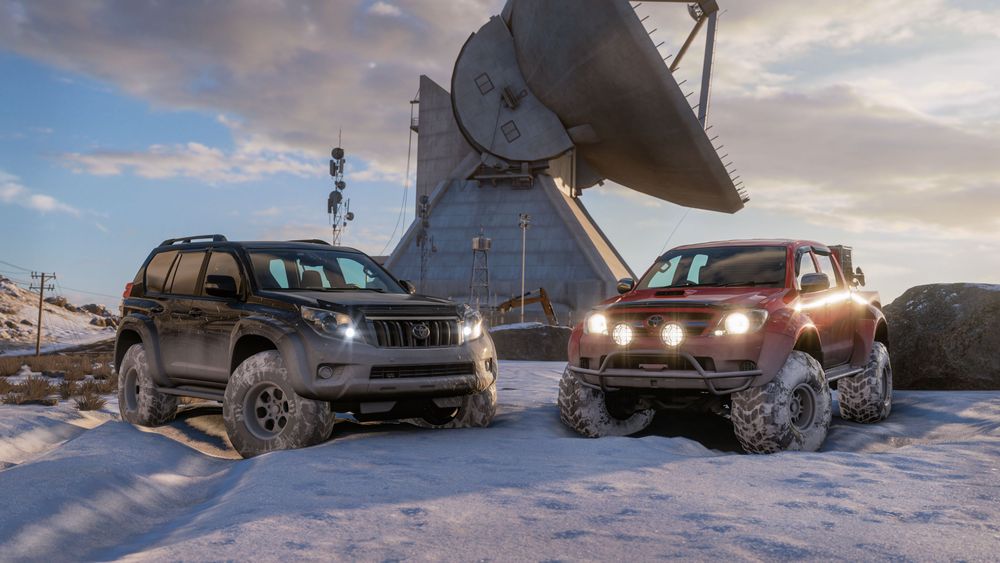 Two Toyota Arctic trucks, one black, the other red, are parked in the snow in front of a large satellite dish.