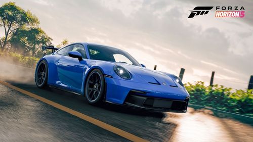 A blue Porsche 911 GT3 speeds down the road with reflections casted by sunlight visible on the road's wet surface.